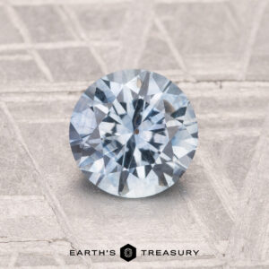 A 1.26-carat heated Montana sapphire, cut in a round brilliant shape, rests on a textured gray surface with "Earth's Treasury" displayed below.