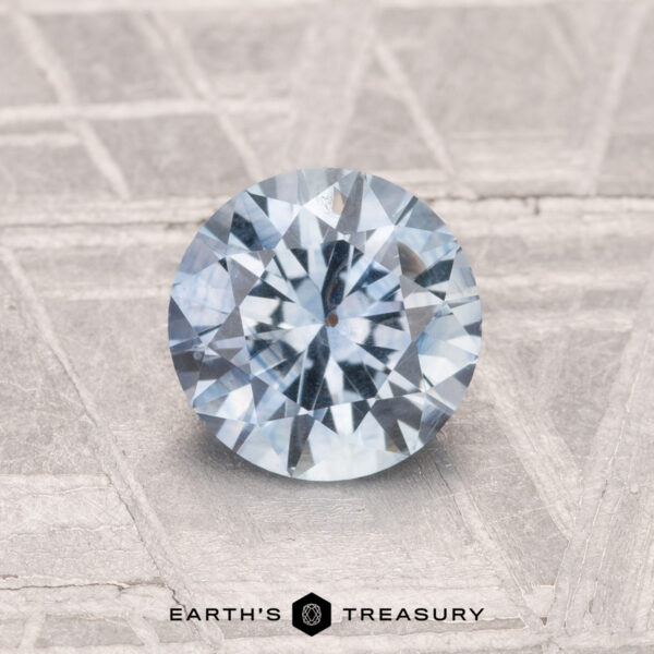 1.26-Carat Montana Sapphire (Heated) A 1.26-carat heated Montana sapphire, cut in a round brilliant shape, rests on a textured gray surface with "Earth's Treasury" displayed below.