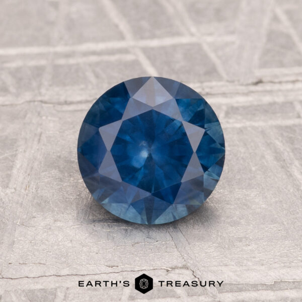 1.26-Carat Montana Sapphire (Heated) A 1.26-carat heated Montana sapphire, round and faceted, is shown on a textured gray surface with "Earth's Treasury" at the bottom.