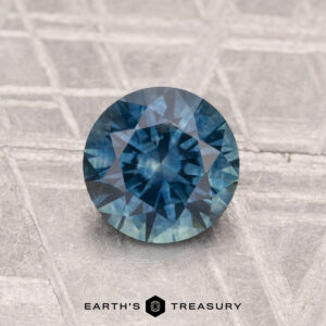 A 1.20-carat heated Montana sapphire, cut in a round brilliant shape, is shown on a textured gray surface with "EARTH'S TREASURY" text at the bottom.