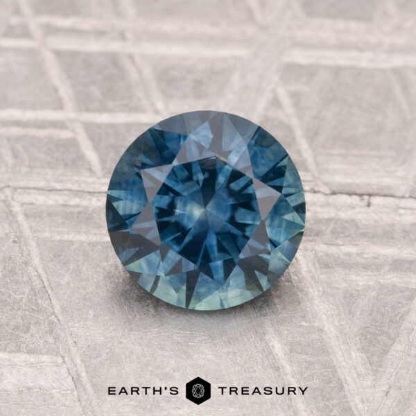1.20-Carat Montana Sapphire (Heated) A 1.20-carat heated Montana sapphire, cut in a round brilliant shape, is shown on a textured gray surface with "EARTH'S TREASURY" text at the bottom.