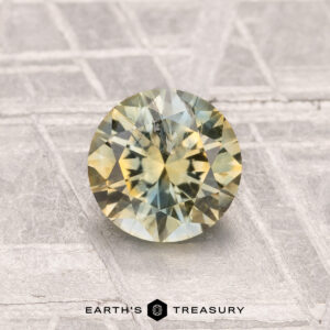 A 1.16-carat heated Montana sapphire with yellow-green hues is displayed on a textured gray surface, featuring the text "EARTH'S TREASURY" at the bottom.