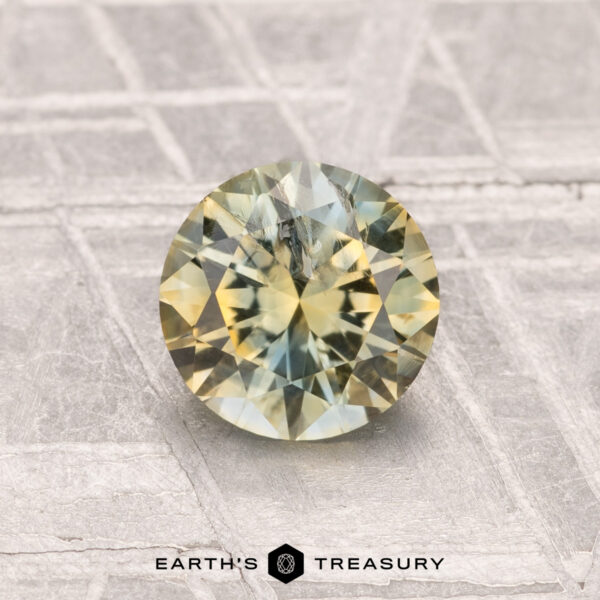 1.16-Carat Montana Sapphire (Heated) A 1.16-carat heated Montana sapphire with yellow-green hues is displayed on a textured gray surface, featuring the text "EARTH'S TREASURY" at the bottom.