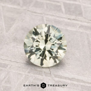 A 1.12-carat round-cut pale yellow Montana sapphire (heated) rests on a textured light gray surface with "Earth's Treasury" below.