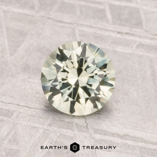 1.12-Carat Montana Sapphire (Heated) A 1.12-carat round-cut pale yellow Montana sapphire (heated) rests on a textured light gray surface with "Earth's Treasury" below.