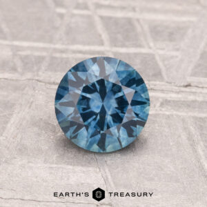 A 1.01-carat heated Montana sapphire with a round brilliant cut is displayed on a textured gray surface, with "EARTH'S TREASURY" shown at the bottom.