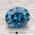 A faceted 3.74-carat heated Montana sapphire with a starburst pattern sits on a textured, light surface. "EARTH'S TREASURY" is displayed at the bottom.