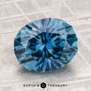 A faceted 3.74-carat heated Montana sapphire with a starburst pattern sits on a textured, light surface. "EARTH'S TREASURY" is displayed at the bottom.