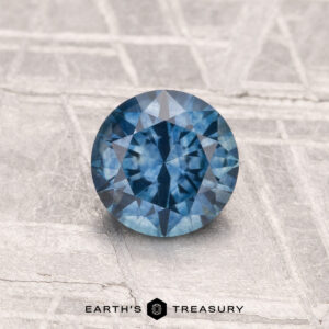 A 0.94-carat heated Montana sapphire, round brilliant-cut and blue, is displayed on a textured gray surface with "EARTH'S TREASURY" at the bottom.