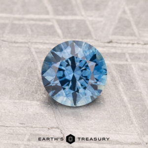A 0.94-carat heated Montana sapphire with a brilliant round cut rests on a textured gray surface. "EARTH'S TREASURY" appears at the bottom of the image.