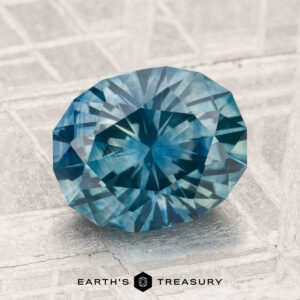 A 3.18-carat heated Montana sapphire, round and multi-faceted with a vivid blue hue, is displayed on a light textured surface above the text "EARTH'S TREASURY.