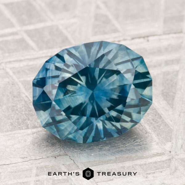 3.18-Carat Montana Sapphire (Heated) A 3.18-carat heated Montana sapphire, round and multi-faceted with a vivid blue hue, is displayed on a light textured surface above the text "EARTH'S TREASURY.