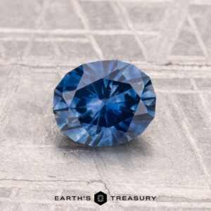 A 1.22-carat heated Montana sapphire, faceted in an oval shape, is displayed on a metallic textured surface with "EARTH'S TREASURY" at the bottom.