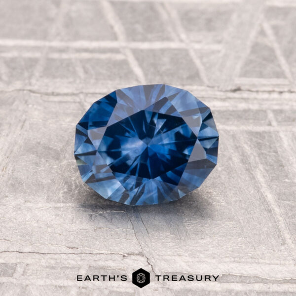 A 1.22-carat heated Montana sapphire, faceted in an oval shape, is displayed on a metallic textured surface with "EARTH'S TREASURY" at the bottom.