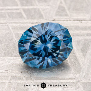 A faceted blue 2.35-carat Montana sapphire (heated) with a starburst pattern rests on a textured surface; "EARTH'S TREASURY" appears at the bottom.
