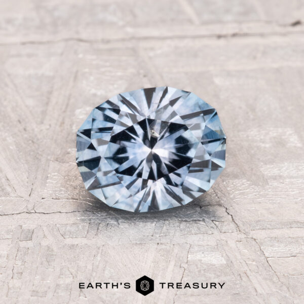 1.07-Carat Montana Sapphire (Heated) A 1.07-Carat Montana Sapphire (Heated) with a blue-gray hue rests on a textured light gray surface, with the words "EARTH'S TREASURY" visible at the bottom.