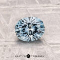 A 1.07-carat heated Montana Sapphire in aqua blue, round brilliant-cut, is displayed on a textured gray surface with the "EARTH'S TREASURY" logo at the bottom.