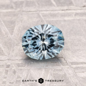 A 1.07-carat heated Montana Sapphire in aqua blue, round brilliant-cut, is displayed on a textured gray surface with the "EARTH'S TREASURY" logo at the bottom.