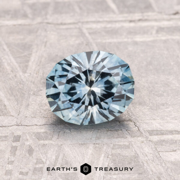 1.07-Carat Montana Sapphire (Heated) A 1.07-carat heated Montana Sapphire in aqua blue, round brilliant-cut, is displayed on a textured gray surface with the "EARTH'S TREASURY" logo at the bottom.