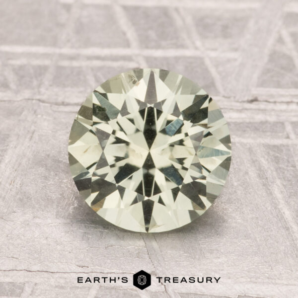 1.81-Carat Montana Sapphire (Heated) A 1.81-carat heated Montana sapphire, round brilliant-cut with a pale green hue, is displayed on a textured surface with "EARTH'S TREASURY" at the bottom.