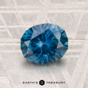 A 1.59-carat heated Montana sapphire with an oval cut is displayed on a textured gray surface, with "EARTH'S TREASURY" shown below the gemstone.