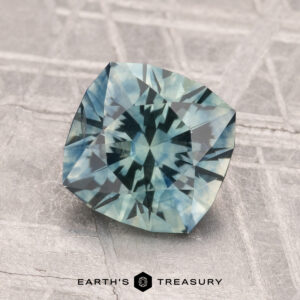 A faceted blue-green 2.18-carat Montana Sapphire (Heated) with a geometric cut rests on a textured gray surface; “EARTH'S TREASURY” is printed below the gem.