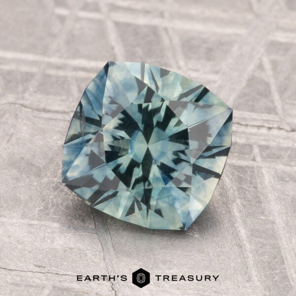 2.18-Carat Montana Sapphire (Heated) A faceted blue-green 2.18-carat Montana Sapphire (Heated) with a geometric cut rests on a textured gray surface; “EARTH'S TREASURY” is printed below the gem.