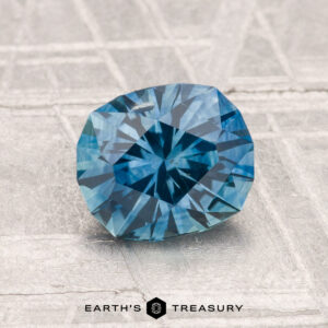A 1.54-carat heated Montana sapphire in a round brilliant cut rests on a textured light gray surface, with "EARTH'S TREASURY" featured at the bottom.