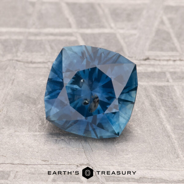 1.62-Carat Montana Sapphire (Heated) A 1.62-carat heated Montana Sapphire with visible inclusions and a cushion cut rests on a textured gray surface, with the "Earth's Treasury" logo displayed at the bottom of the image.