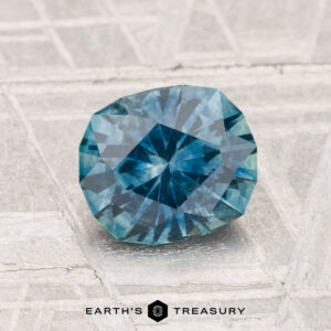 A 1.51-carat heated Montana sapphire rests on a textured metallic surface with "EARTH'S TREASURY" displayed at the bottom.