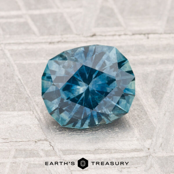 A 1.51-carat heated Montana sapphire rests on a textured metallic surface with "EARTH'S TREASURY" displayed at the bottom.