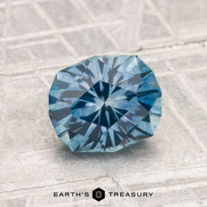 A 1.76-carat heated Montana sapphire is displayed on a textured gray surface, with the "EARTH'S TREASURY" logo at the bottom.