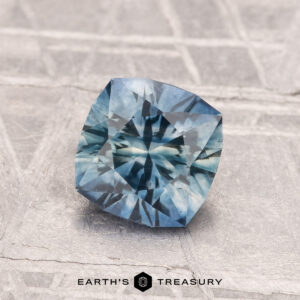 A 1.30-carat cushion-cut heated Montana sapphire with intricate facets is displayed on a textured gray surface. The "Earth's Treasury" logo appears at the bottom.