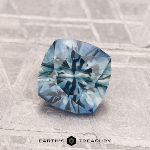 1.30-Carat Montana Sapphire (Heated) A 1.30-carat cushion-cut heated Montana sapphire with intricate facets is displayed on a textured gray surface. The "Earth's Treasury" logo appears at the bottom.