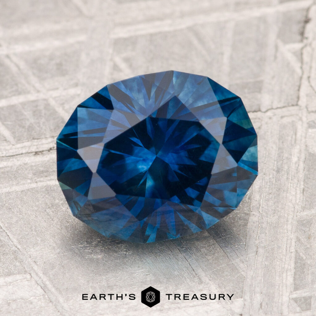 A faceted deep blue 2.96-carat heated Montana sapphire rests on a textured metallic surface with the "EARTH'S TREASURY" logo and text below.