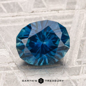 A faceted round blue 2.85-carat Montana Sapphire (Heated) rests on a textured gray surface, with the text “Earth’s Treasury” at the bottom.