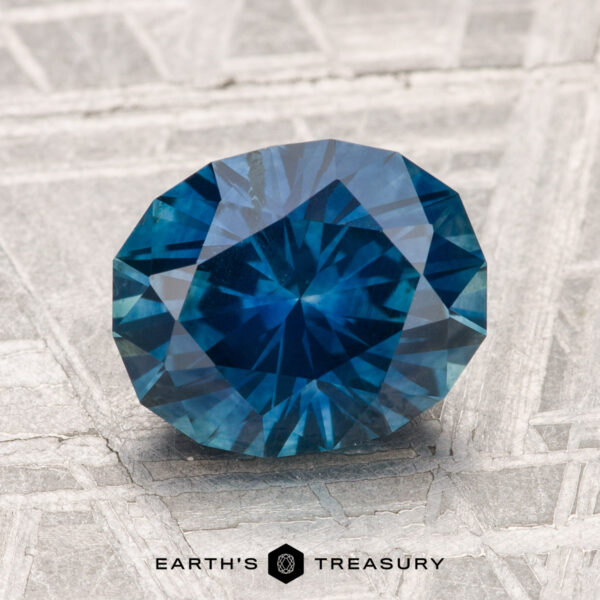 2.85-Carat Montana Sapphire (Heated) A faceted round blue 2.85-carat Montana Sapphire (Heated) rests on a textured gray surface, with the text “Earth’s Treasury” at the bottom.