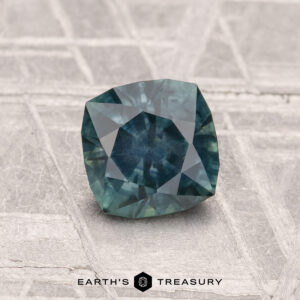 A faceted 1.08-carat heated Montana sapphire in blue-green rests on a textured surface, with "EARTH’S TREASURY" and its logo visible at the bottom of the image.
