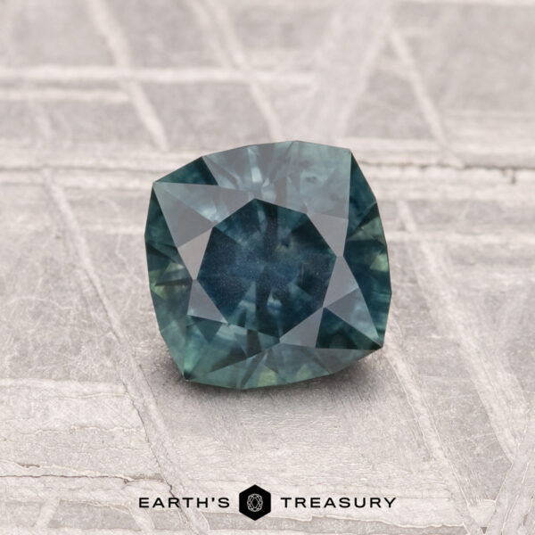 1.08-Carat Montana Sapphire (Heated) A faceted 1.08-carat heated Montana sapphire in blue-green rests on a textured surface, with "EARTH’S TREASURY" and its logo visible at the bottom of the image.