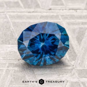 A 2.23-carat heated Montana sapphire with a deep blue hue is showcased on a textured, light-toned surface, labeled "Earth's Treasury" at the bottom.