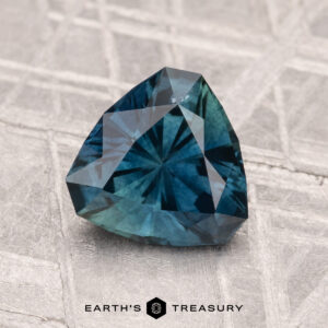 A faceted, trillion-cut 1.58-carat heated Montana sapphire in blue-green rests on a textured gray surface. "EARTH'S TREASURY" appears at the bottom.
