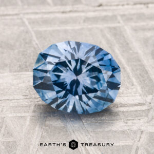 A 1.94-carat heated Montana sapphire is displayed on a textured surface with the text "EARTH'S TREASURE" and a logo below it.
