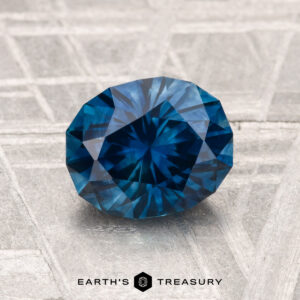 A 1.75-carat heated Montana sapphire, cut in a faceted oval shape, is displayed on a textured light-colored surface with "EARTH'S TREASURY" below.