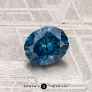 A faceted 1.16-Carat Montana Sapphire (Heated) with intricate cuts rests on a textured metallic surface; "EARTH'S TREASURY" is written at the bottom.