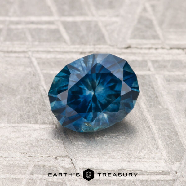 A faceted 1.16-Carat Montana Sapphire (Heated) with intricate cuts rests on a textured metallic surface; "EARTH'S TREASURY" is written at the bottom.