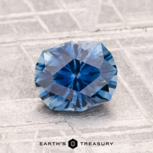 A round, faceted 1.52-carat Montana Sapphire (Heated) sparkles on a textured, light-colored surface. Text at the bottom reads "EARTH'S TREASURY.