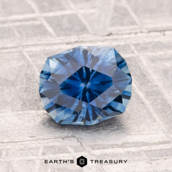1.52-Carat Montana Sapphire (Heated) A round, faceted 1.52-carat Montana Sapphire (Heated) sparkles on a textured, light-colored surface. Text at the bottom reads "EARTH'S TREASURY.