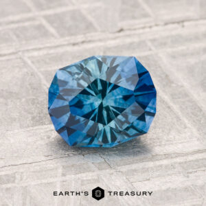 A 1.55-carat heated Montana sapphire with intricate geometric facets rests on a textured light gray surface. "EARTH'S TREASURY" is displayed at the bottom of the image.