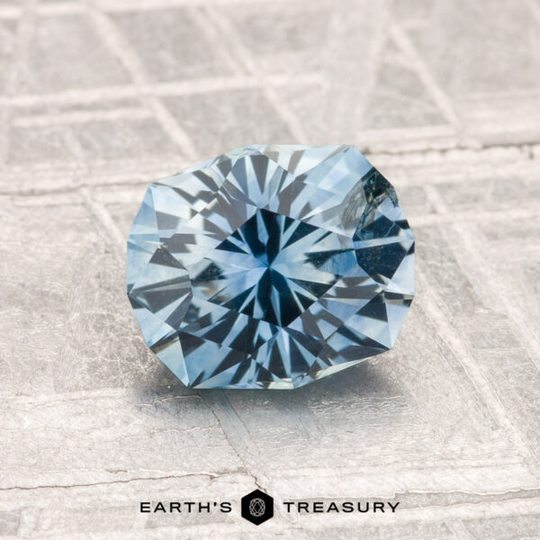1.55-Carat Montana Sapphire (Heated) A round, faceted 1.55-carat heated Montana sapphire is displayed on a textured, light-colored surface with "EARTH'S TREASURY" beneath it.