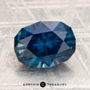 A 3.39-carat heated Montana sapphire with a deep blue color is displayed on a textured light gray surface, with the "Earth's Treasury" logo below.